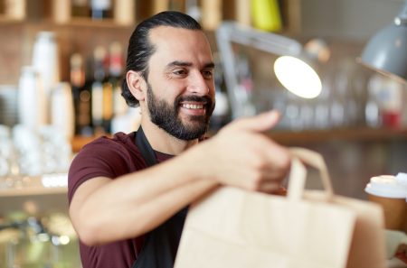 small business, people, takeaway and service concept - happy man or waiter in apron holding coffee cups and paper bag at bar